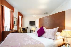 Modern open plan appartment. Bed in the foreground with bedside tables with lamps on, on either side. Desk and sofas toward the back of the room with the television mounted on the far war.
